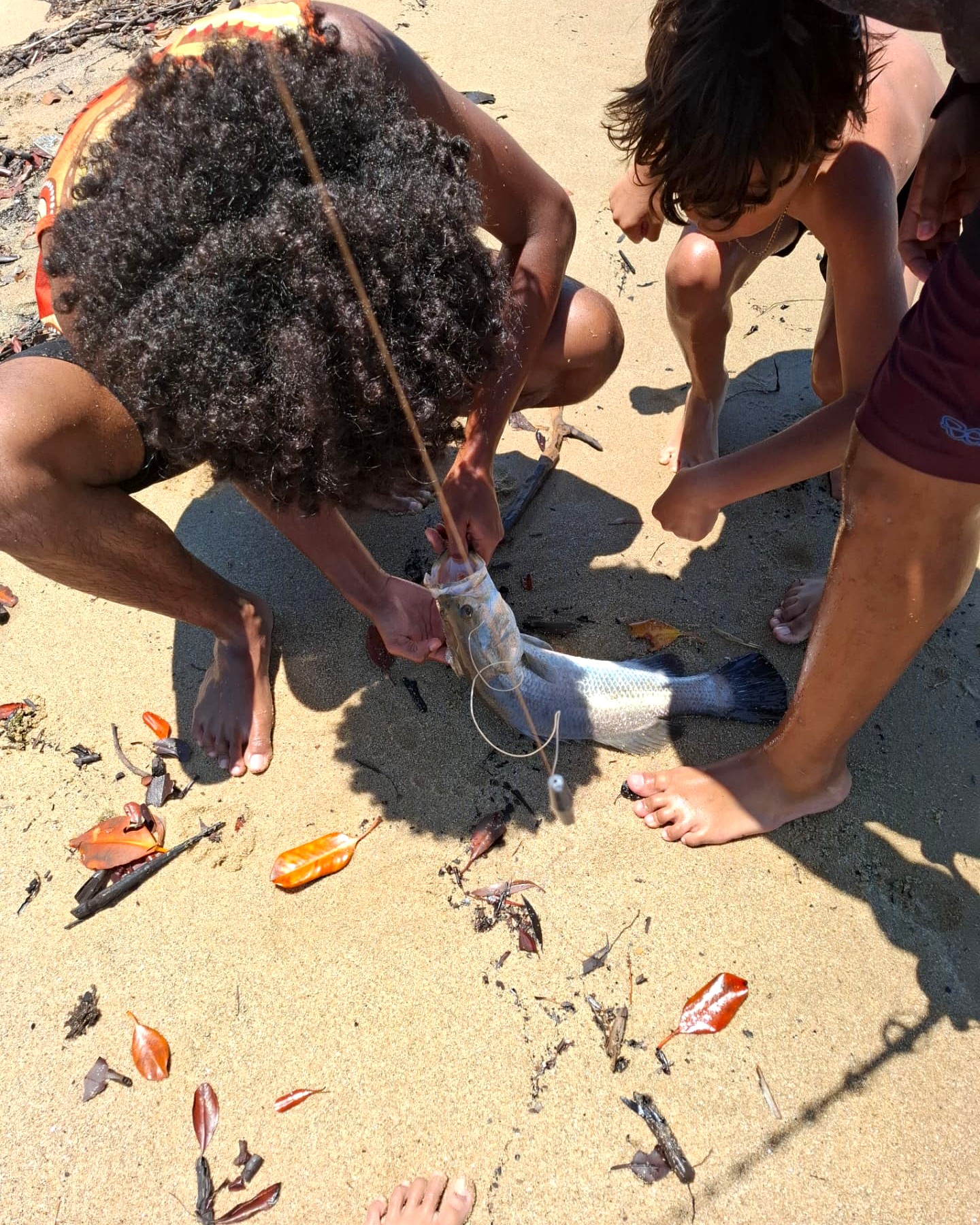 Young people gathered around a freshly caught fish on a sandy shoreline.