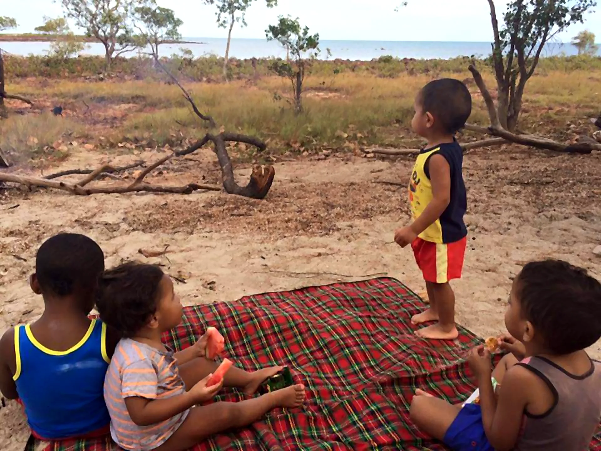 Four children sharing watermelon on a picnic mat beside the water.